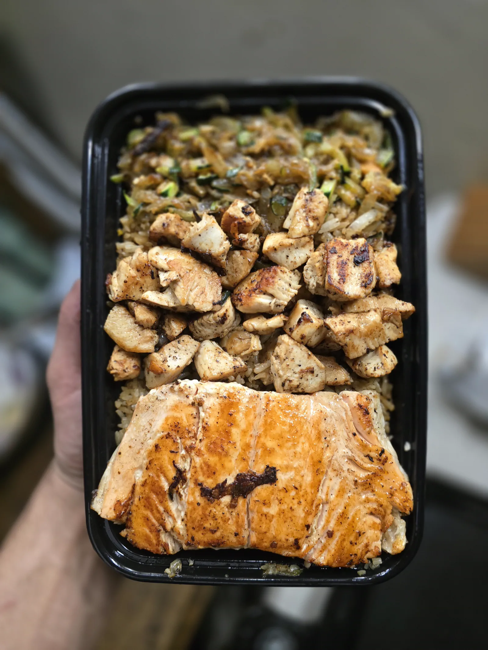 Seared chicken and a whole salmon fillet over garlic fried rice, held up in a black tray