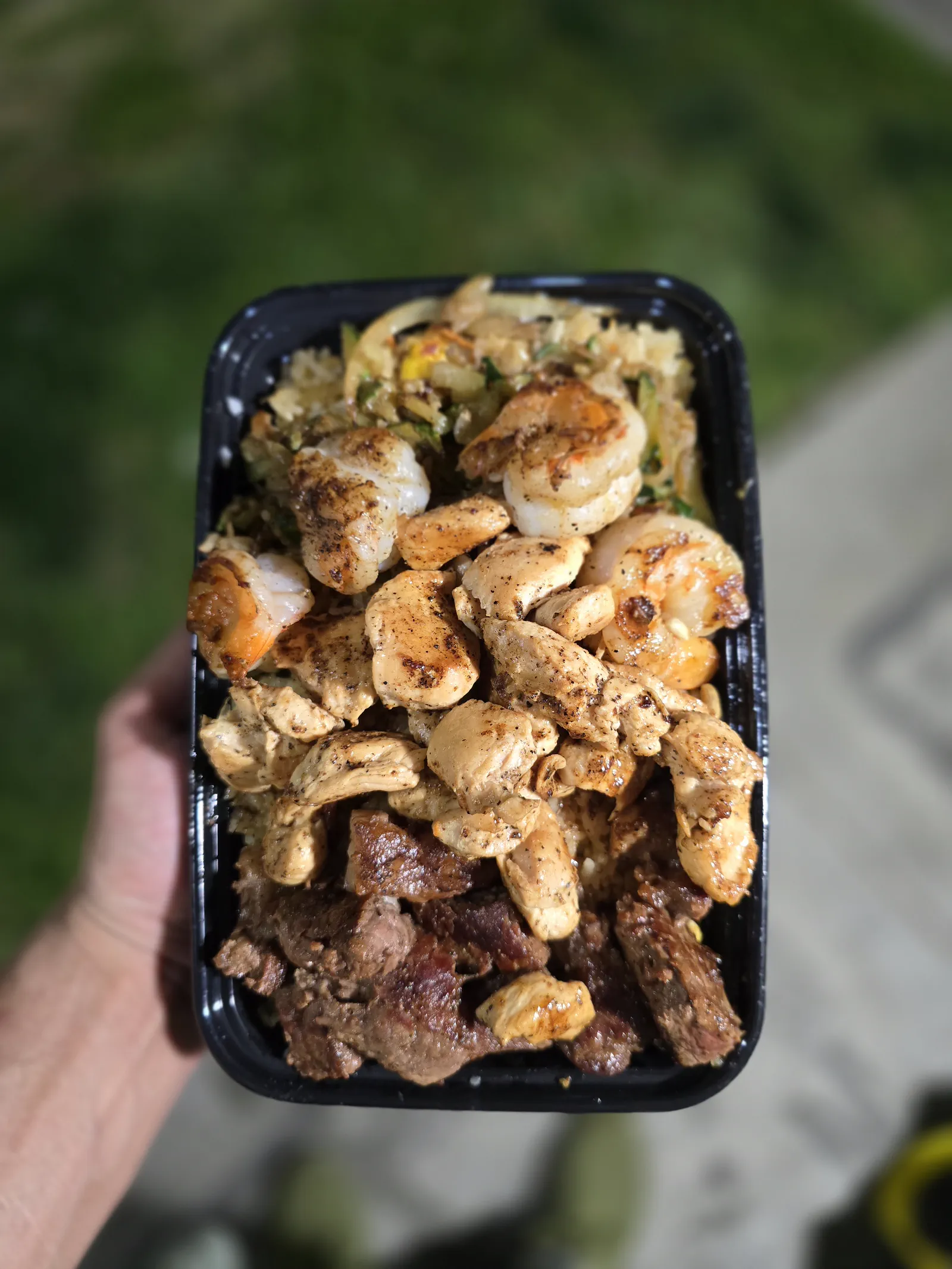 Seared shrimp, chicken and steak piled over garlic fried rice, held up outdoors in a black tray
