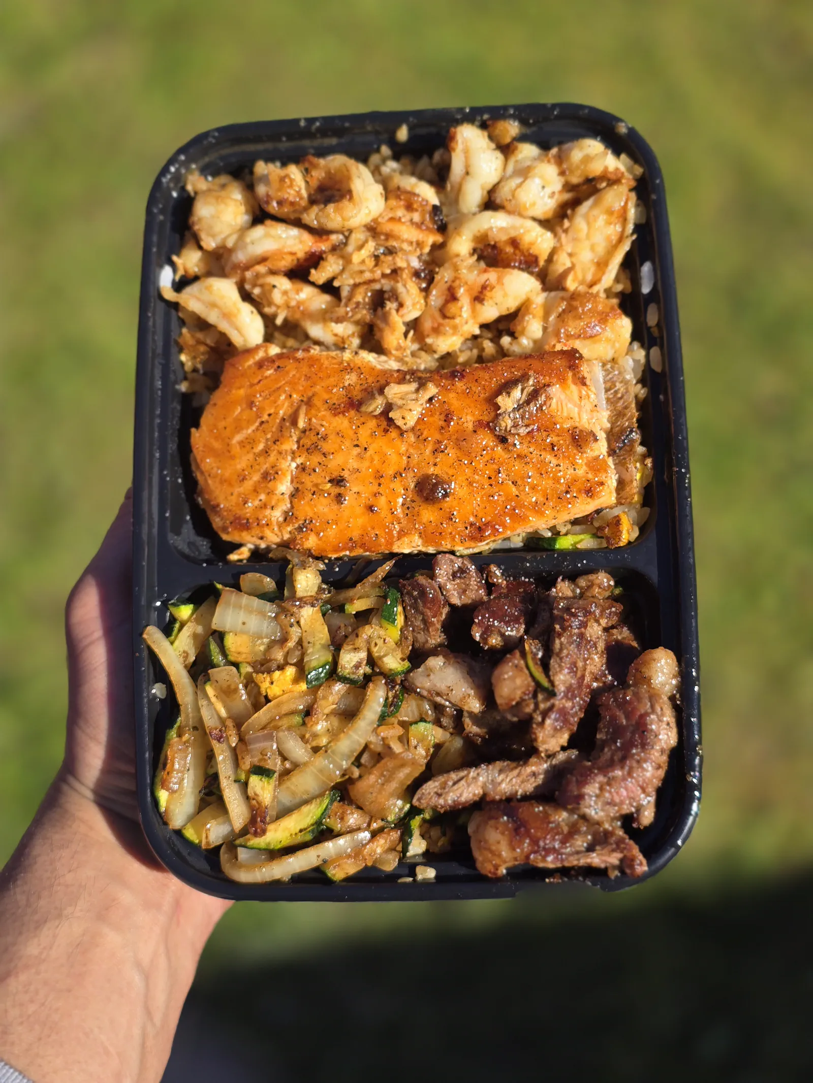 Seared shrimp, blackened salmon fillet and steak with zucchini over garlic fried rice, held up outdoors in a two-compartment black tray